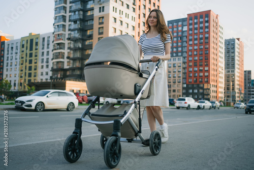 Young woman pushing a baby stroller along a city street, engaging in daily life and urban motherhood with high rise residential buildings in the background