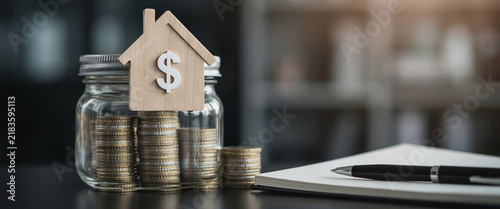 Glass jar with money coins and house shaped wooden symbol for savings growth plan.
