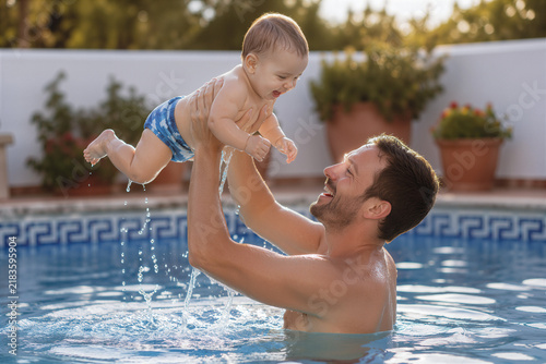Father lifting baby high in sunny outdoor swimming pool