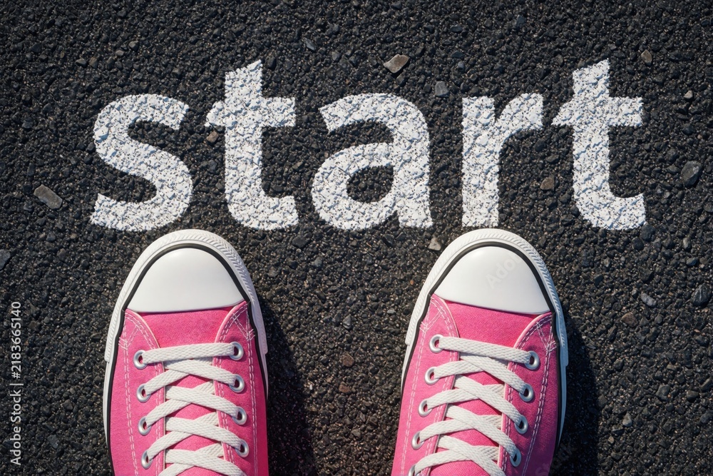 Obraz premium Top view of pink sneakers on black asphalt road. White text says start on ground. Woman stands at starting line ready to begin journey race. Concept of new beginning, motivation, personal goal career