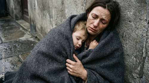 Fototapeta kuchenna A distressed woman embraces a sleeping child, both wrapped in a blanket, seeking comfort against a cold wall.