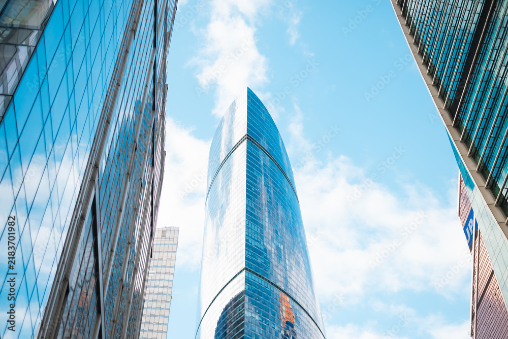 Obraz premium A tall, modern skyscraper stands between two glass buildings in an urban setting. The structure reflects the clear blue sky and scattered clouds, showcasing contemporary architectural designs