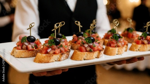 Waiter holds white platter with gourmet bruschetta appetizers. Italian crostini topped with tomato, onion, herbs, and olive. Ideal for events, weddings, parties, catering.