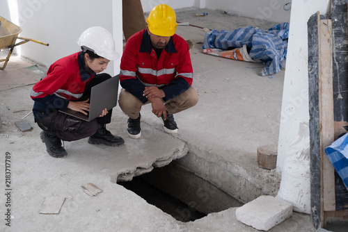 Construction engineers inspecting an open concrete floor trench with a laptop during building site evaluation, highlighting structural inspection, workplace safety, foundation assessment, teamwork.