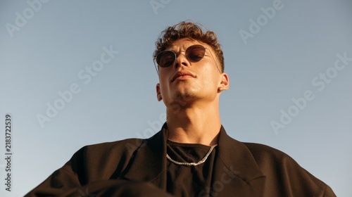 Young Man with Tattoo in Urban Setting, Casual Streetwear Fashion, Sunglasses, Necklace, Blue Jeans, Graffiti Background