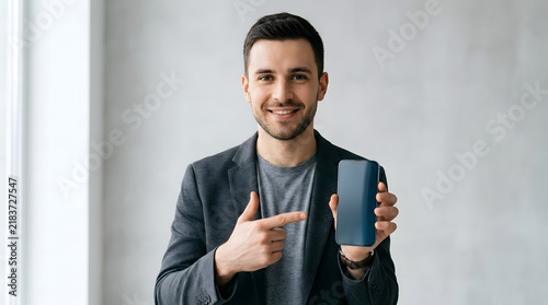 Smiling man in blazer pointing at blank smartphone screen for mobile app presentation