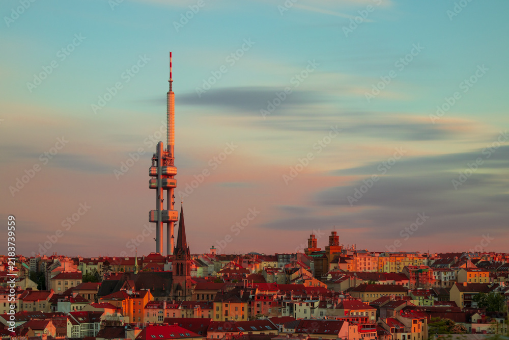 Obraz premium Žižkov Television Tower rising above Prague at sunset, with St. Procopius Church and red-roofed cityscape glowing in warm evening light, Czech Republic.