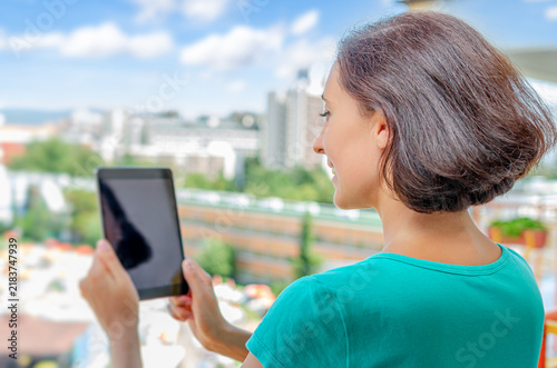 A woman stands outdoors on a sunny day while using a digital tablet with a city background.