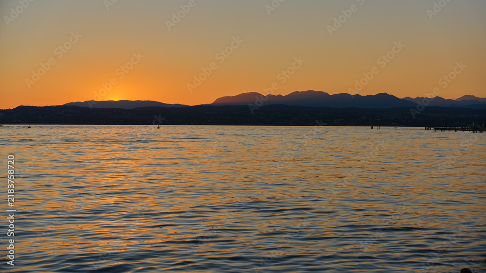 Obraz premium Wide panoramic view of Lake Garda at dusk with warm orange sunset glow illuminating water ripples and distant alpine mountain ridges. Concept of tranquil evening waterscape, Italian lakes vacation, sc
