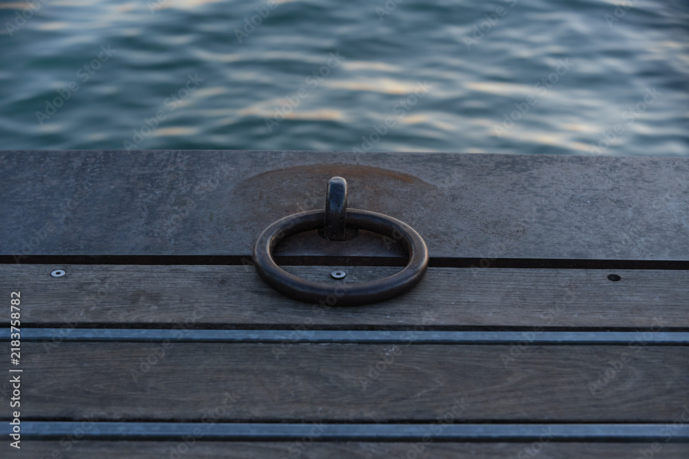 Obraz premium Close-up of a sturdy steel mooring ring on a wooden dock with calm water ripples in the background at Lake Garda. Concept of maritime equipment, boat mooring, nautical transport, pier construction, an