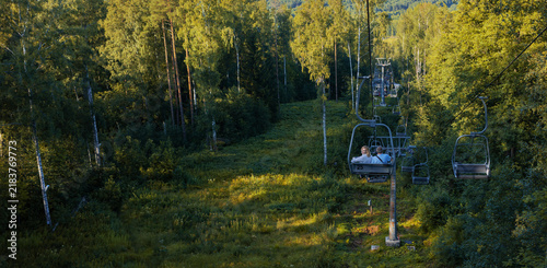 Golden evening light on a forest slope, chairlift carrying a couple above a grassy hillside.