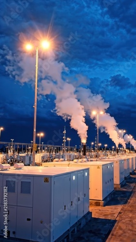 Industrial power generation facility at dusk with illuminated equipment and steam emissions rising against a dramatic blue sky filled with clouds