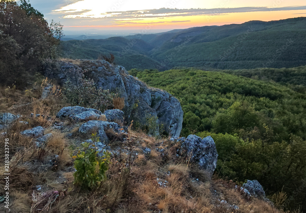 Fototapeta premium Rugged limestone rocks and dry grass on Kopus-ko overlooking the densely forested Garadna Valley at dawn in the Bukk Mountains, Hungary.