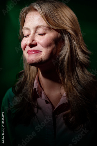 A close-up portrait of a woman smiling with her eyes closed, conveying calm joy and contentment. Natural freckles, soft lighting and relaxed expression create an intimate, candid emotional moment.