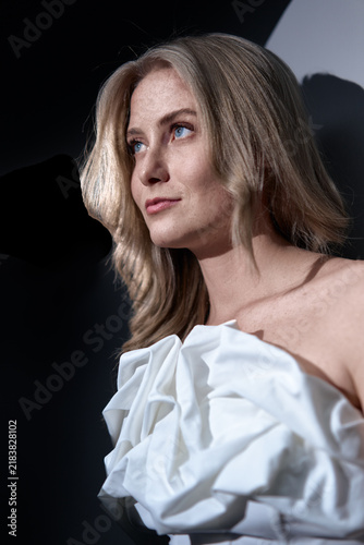 Картината върху платно Elegant blonde woman gazes upward in a dramatic studio portrait wearing a white ruffled dress