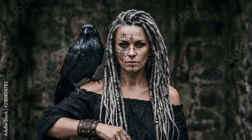 A fierce viking woman with dreadlocks and war paint standing with a black raven on her shoulder