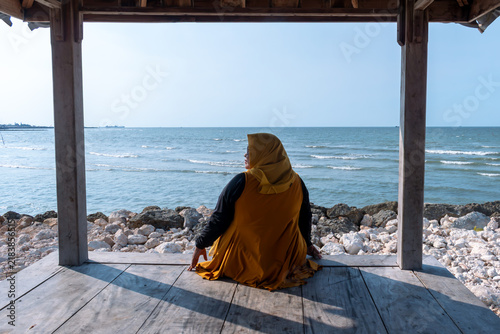 Obraz na plátně Woman wearing yellow hijab looks at distant sea from wooden gazebo for quiet solitude