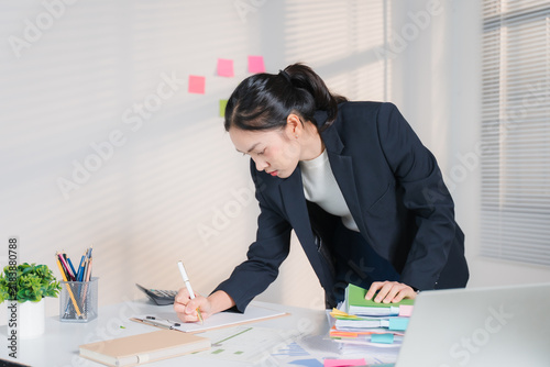 Quadro su tela Focused Female Professional at Work: A dedicated businesswoman in a smart suit leans over a desk, meticulously reviewing documents and making notes
