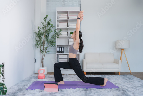 Yoga Practice at Home: A woman in athletic wear performs a warrior pose on a yoga mat in a bright, airy living room, guided by a tablet, promoting wellness and a healthy lifestyle.