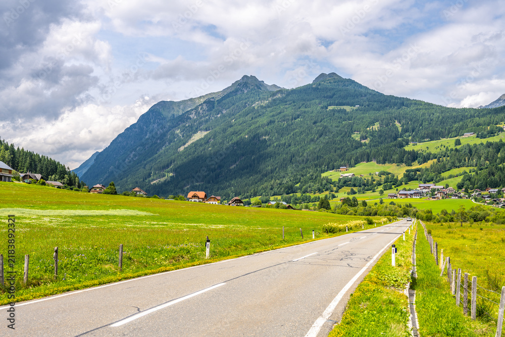Obraz premium A winding alpine road stretches through green fields with mountains in the background. Houses dot the landscape under a cloudy sky. The area is vibrant with nature's colors.