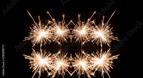 Symmetrical composition of four burning hand-held sparklers with glowing golden sparks reflecting on a dark water-like surface against a solid black background.