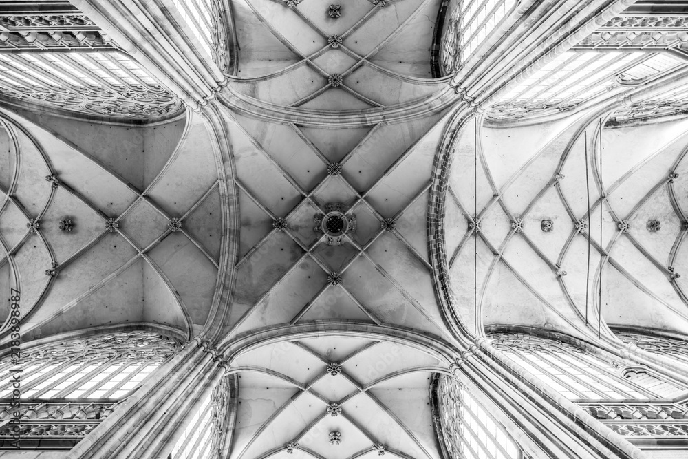 Obraz premium Visitors look up at the ceiling of St Vitus Cathedral in Prague. The design features arches and windows. Sunlight shines through the glass and illuminates the stone structure.