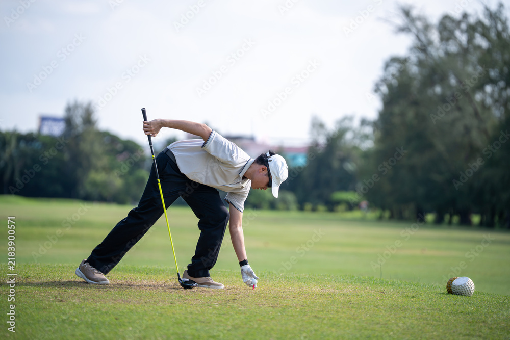 Obraz premium Asian Golfer Placing a Golf Ball on a Tee While Holding a Driver and Preparing for a Focused Tee Shot on a Sunny Outdoor Golf Course