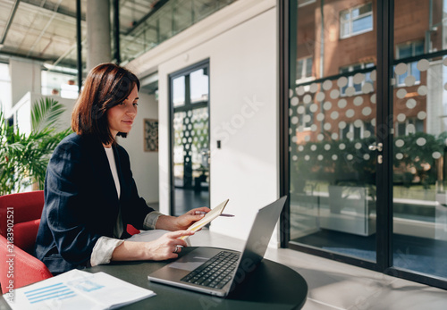 Businesswoman taking notes beside a laptop in a glass office