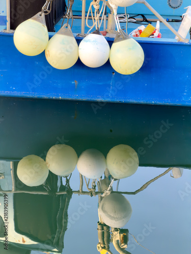 Cream and white fenders hang from a blue boat's side, mirrored upside-down in still, glassy water below