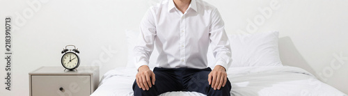 man crisp white shirt dark trousers sits bed minimalist nightstand beside clock table showing soft