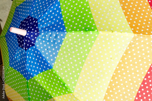 Bright umbrella featuring segmented panels in various colors such as blue, green, yellow, and orange. Each panel is decorated with evenly spaced white polka dots. Umbrella rod is visible, highlighting