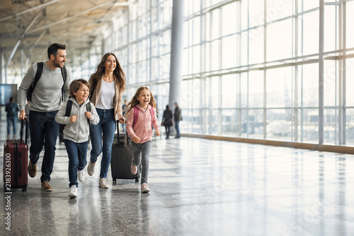 Family Vacation at the Airport: A smiling family, including parents and two children, are pulling their luggage and walking through a modern airport terminal.