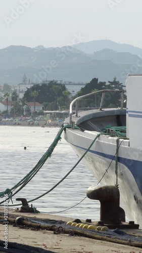 Close up of a boat hull moored with heavy ropes tied to a metal bollard on the dock of a coastal harbor with mountains in the background