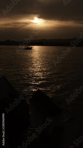 Scenic view of a motorboat cruising across the sea during a golden sunset with sun shining through clouds and rocks in the foreground