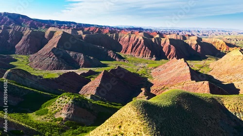 Aerial view of a vast Danxia landform with red sandstone ridges, deep ravines, green vegetation, and bright sunlight beneath a blue sky.
