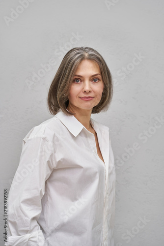 Middle Aged Woman With Short Gray Hair Posing In White Shirt Against Light Gray Studio Background.