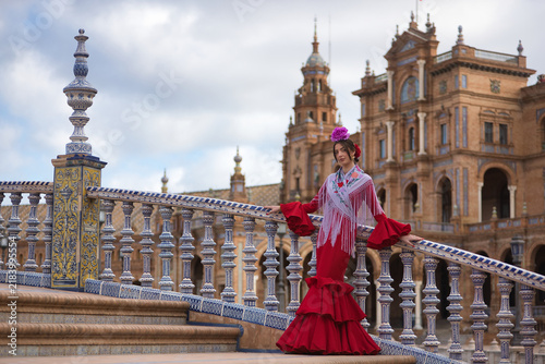 A young Andalusian woman wearing a red flamenco dress with ruffles and a purple shawl, with flowers in her hair, posing while leaning against a beautiful bridge in Spain Square in Seville.