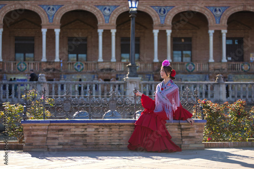 A young woman from Andalusia, wearing a red flamenco dress with ruffles and a purple shawl, posing while seated on a bench in Spain Square in Seville. Concept: dance, art.