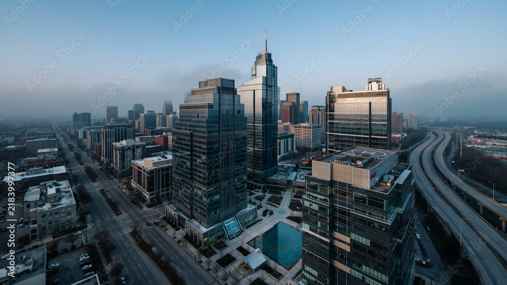 Obraz premium Aerial view of a city skyline with tall glass skyscrapers, office buildings, and a highway during a foggy morning with soft blue lighting