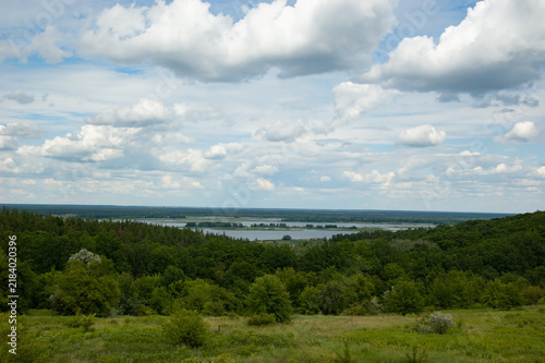 Summer landscape with a grassy meadow, forest, and the Dnipro River with islands, distant shore, blue sky and fluffy white clouds.