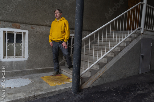 A 40-year-old man in a yellow hoodie stands on outdoor concrete stairs with white railings next to a city building.