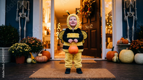 Toddler in bee costume standing on Halloween porch holding pumpkin bucket.