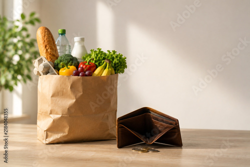 Empty wallet with a few coins next to a paper grocery bag filled with fresh food on a wooden table. Concept of rising food prices, inflation, household expenses and cost of living.