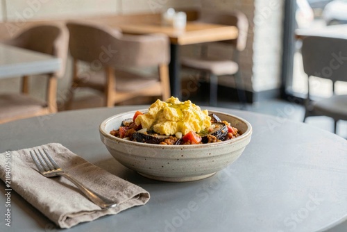 Bowl of Mirza Ghasemi with smoked eggplant, tomato, garlic, and eggs on linen napkin in modern cafe setting. Concept of minimalist design, neutral colors, and tasty dish.