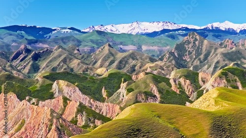 Aerial view of colorful danxia mountains with layered ridges, green vegetation, and snow-covered peaks beneath a clear blue sky in bright sunlight.