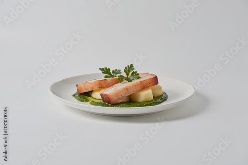 Stegt Flaesk, featuring crisp pork belly topped with parsley sauce. Concept of minimalistic food photography against light grey backdrop, highlighting delicious textures and colors.