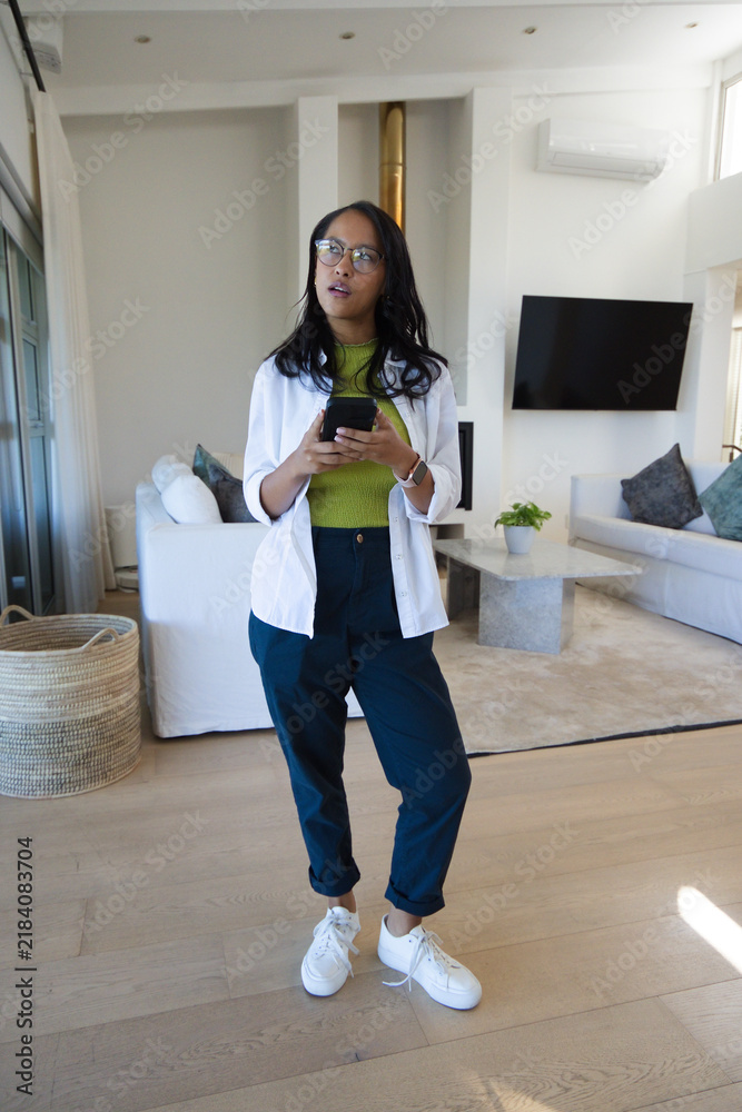 Fototapeta premium Woman standing in center of modern living room holding smartphone and wearing eyeglasses