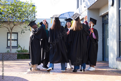 Diverse students wearing black gowns and caps, celebrating in courtyard holding red diploma tubes