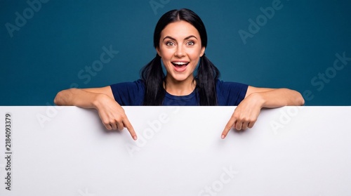 Excited young woman with pigtails pointing down at a large blank white board against blue with index fingers blank