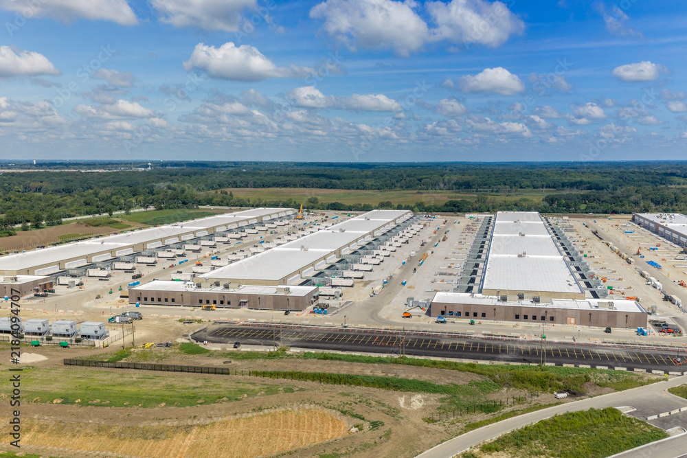 Obraz premium Aerial view of a large data center under construction with expansive industrial buildings, white roofs, parking lots, and surrounding green fields in United States